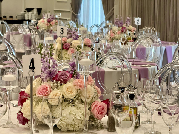 A formal event table decorated with a large floral centerpiece of pink, white, and purple roses, clear acrylic chairs, wine glasses, and table number signs in a banquet hall.