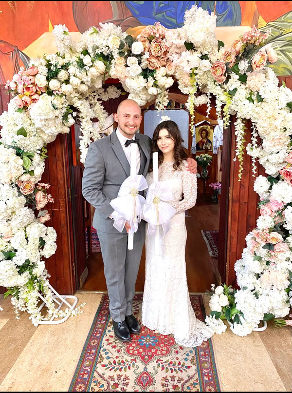A bride and groom standing together under a large floral arch. The groom is wearing a gray suit with a black bow tie, and the bride is wearing a white lace wedding gown.