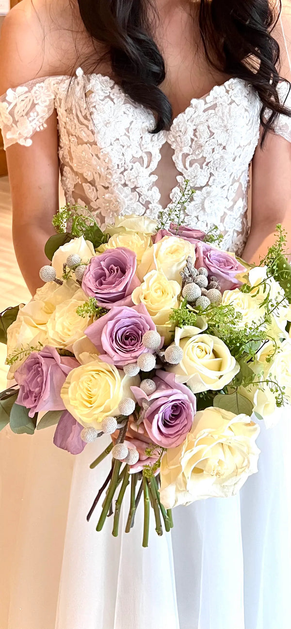 A close-up of a bride in a white lace off-the-shoulder wedding dress holding a large bouquet of lavender and cream roses with green foliage.