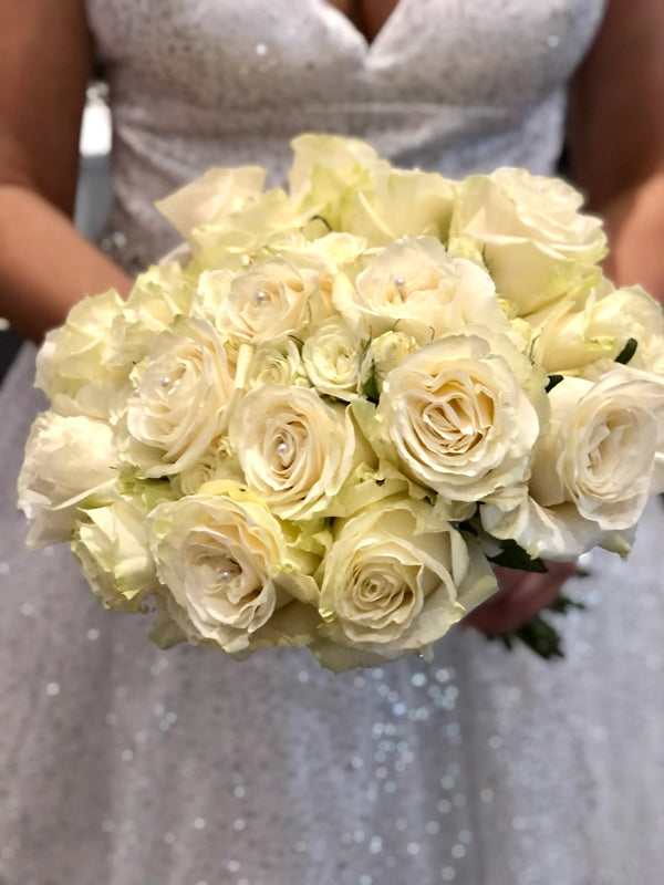 A close-up of a bridal bouquet of cream-colored roses with small pearl accents, held by a person wearing a sparkling silver sequined dress.