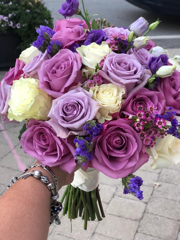 A hand holding a vibrant bouquet of purple and lavender roses, cream lilies, and small purple flowers, with a silver bracelet visible on the wrist, set against an outdoor paved background.