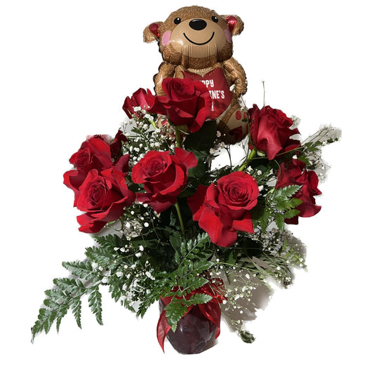 A vase containing a dozen red roses and a decorative bear-shaped balloon, with green foliage around it.