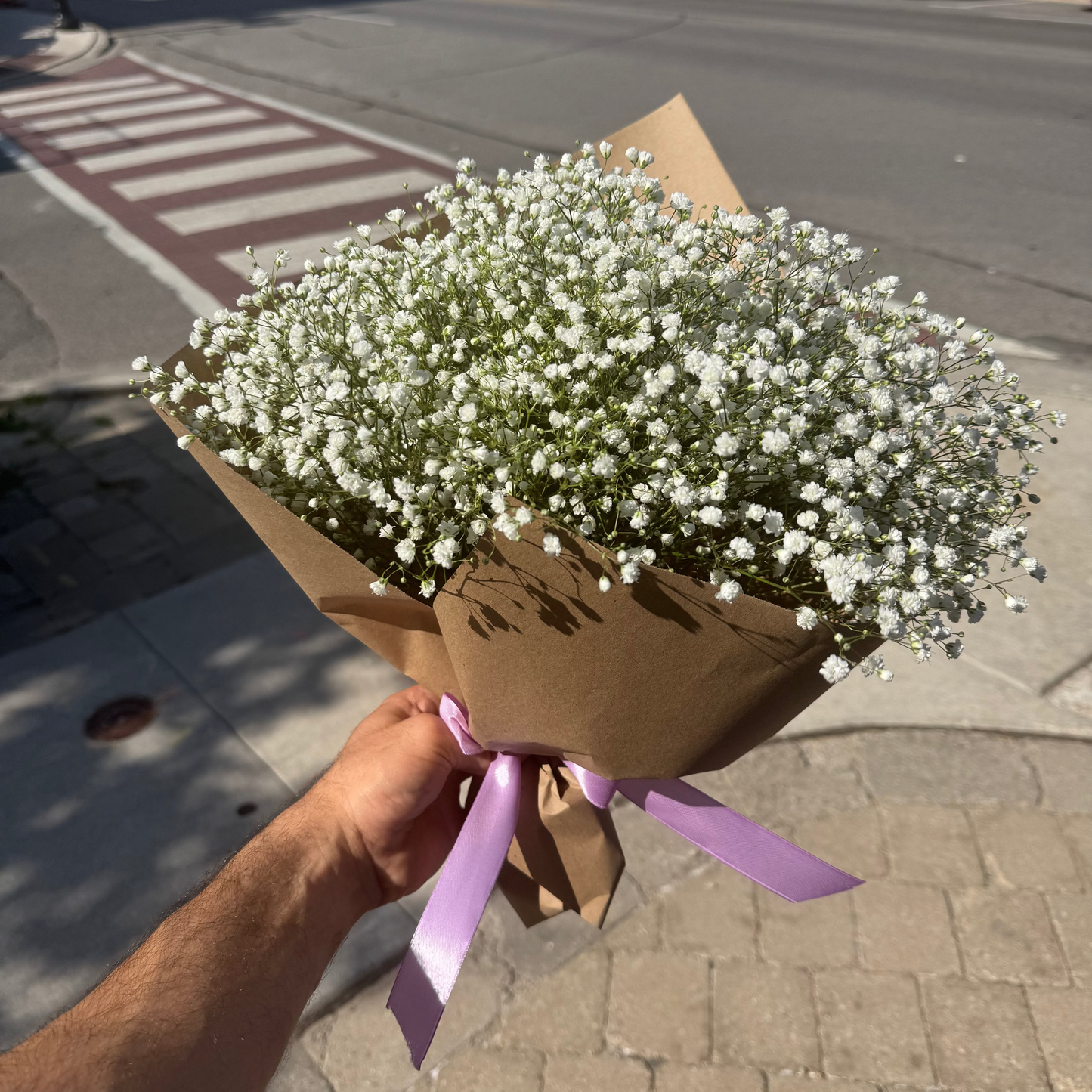 A hand holding a bouquet of white baby's breath flowers wrapped in brown kraft paper with a light purple ribbon, set against a blurred street background.
