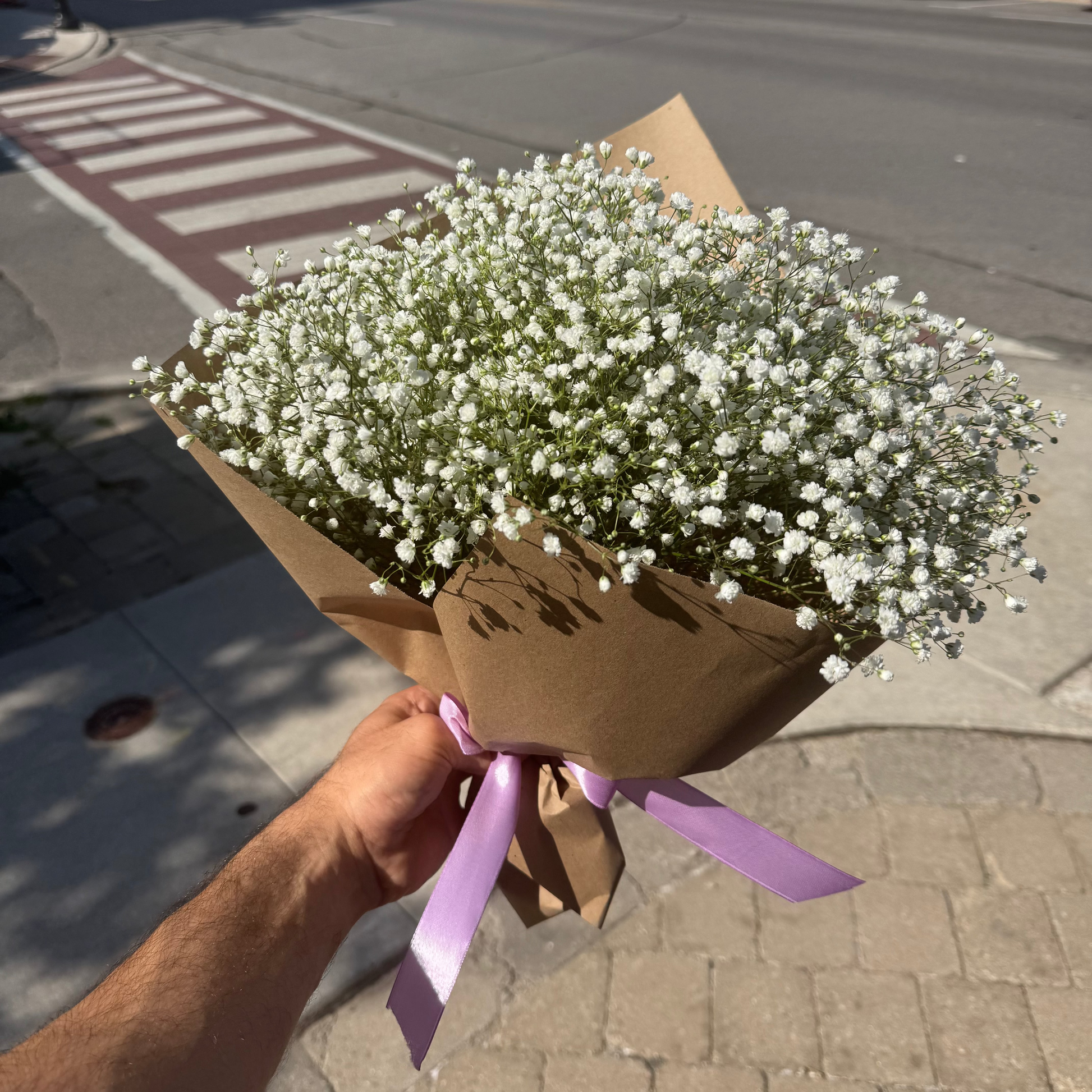 A hand holding a bouquet of white baby's breath flowers wrapped in brown kraft paper with a light purple ribbon, set against a blurred street background.