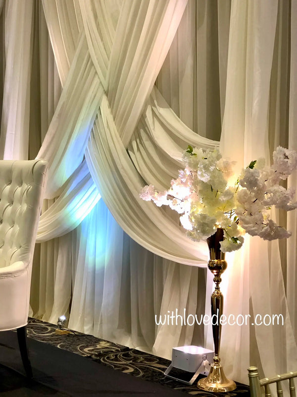 A decorative event backdrop featuring draped cream-colored fabric curtains with soft folds, blue uplighting, a white tufted armchair on the left, and a tall gold floor lamp holding a bouquet of white artificial flowers on the right. A watermark reads 'withlovedecor.