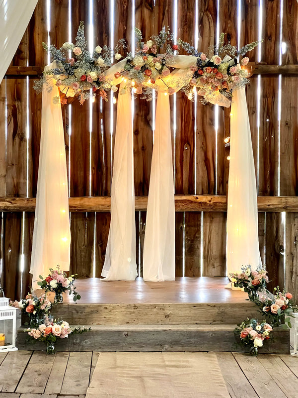 A rustic wedding arch decorated with cream draped fabric, a floral garland with warm string lights, and small flower arrangements on the steps in front, set against a wooden barn wall.