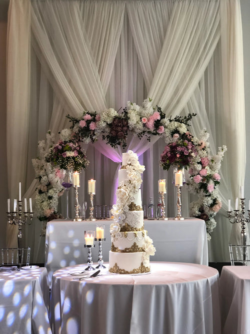A multi-tiered white wedding cake decorated with gold and white floral details, displayed on a round table in front of a draped cream fabric backdrop with a floral arch and candles.