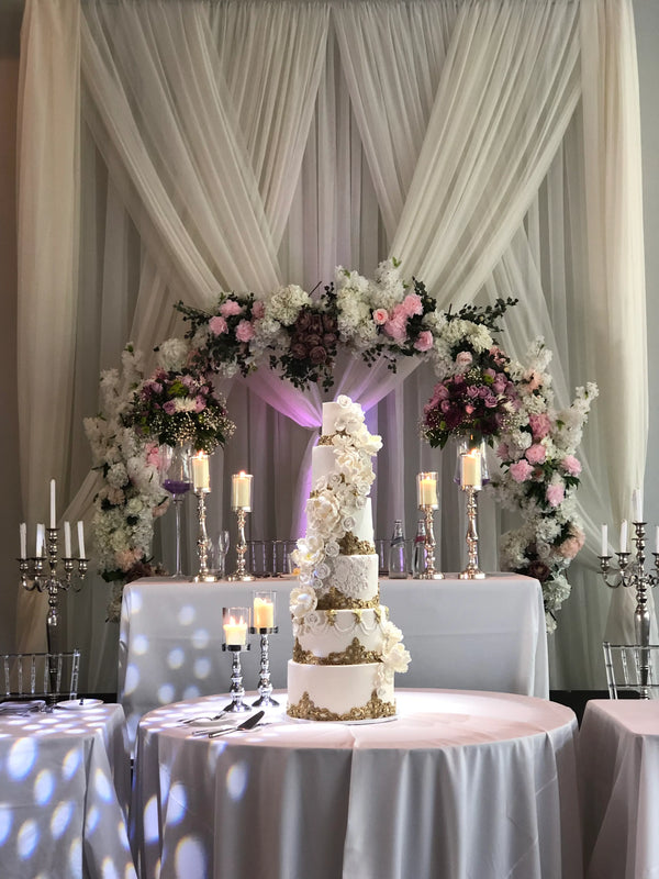 A multi-tiered white wedding cake decorated with gold and white floral details, displayed on a round table in front of a draped cream fabric backdrop with a floral arch and candles.